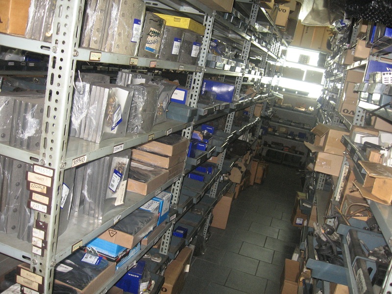 Corridoio di magazzino tra scaffali metallici grigi carichi di ricambi imballati, cartoni e scatole blu Volvo